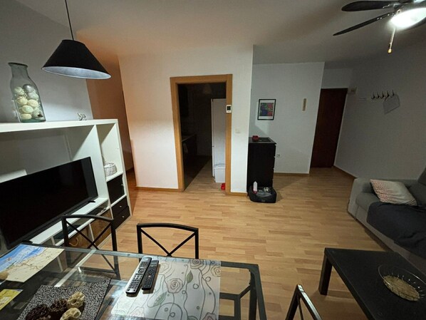 Una sala de estar con un sofá, un televisor, una mesa de comedor con sillas, una mesita de vidrio con controles remotos, un ventilador de techo y un pasillo que lleva a otra habitación.