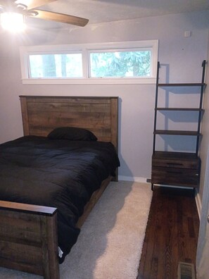 Un dormitorio con cabecera de madera, cama con cubrecama oscuro, alfombra blanca, ventana con vistas a árboles y una escalera de madera apoyada en la pared.