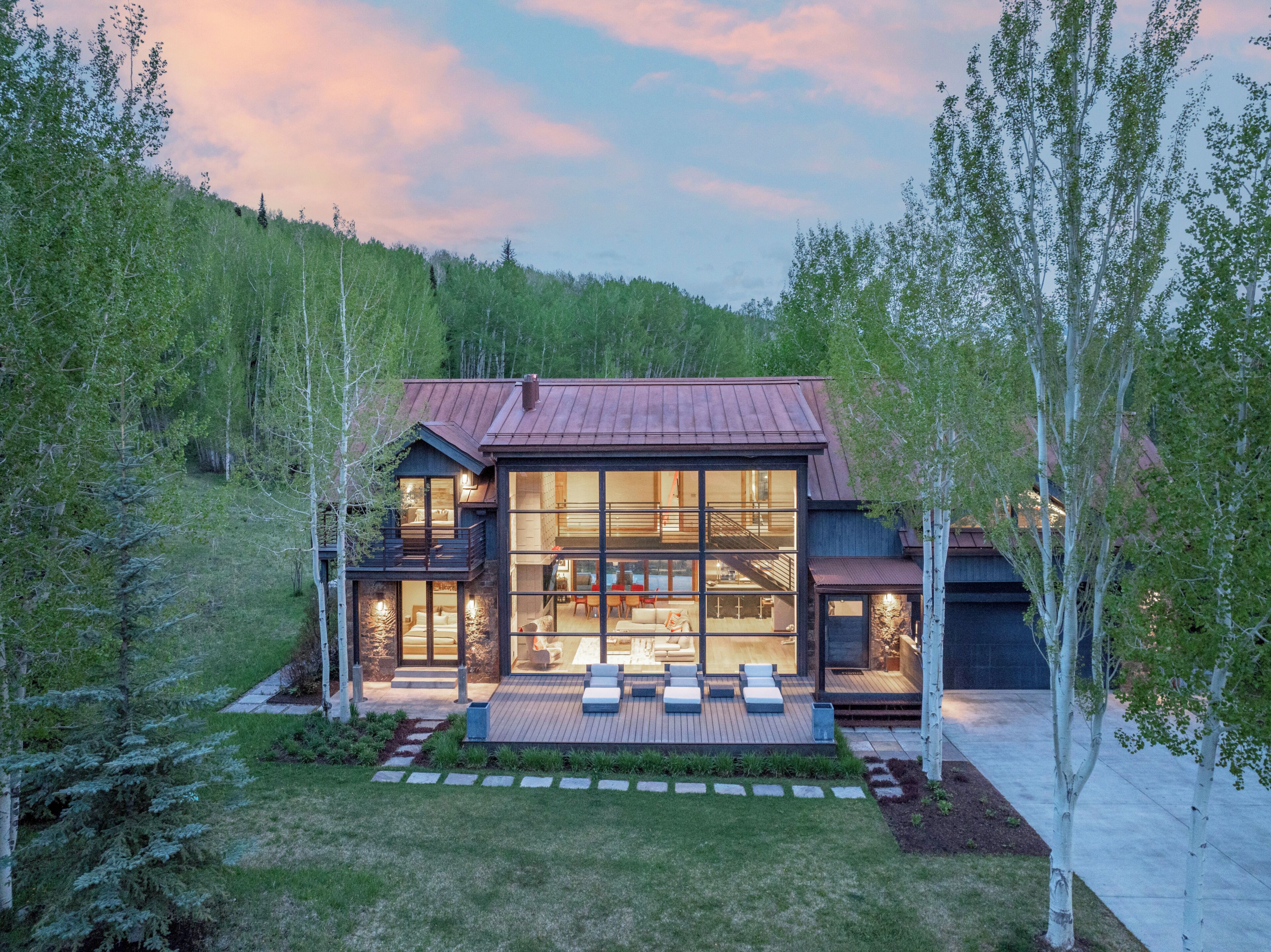 A modern house with a large glass facade, a wooden deck, and a stone pathway leading to the entrance.