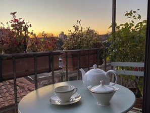 Una mesa con tetera y tazas en un balcón con vista a un paisaje urbano al atardecer.