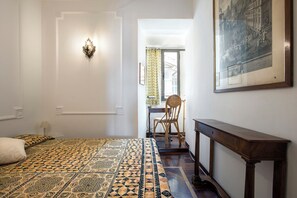 Un dormitorio con una colcha estampada, un escritorio con una silla, una lámpara de pared y un cuadro enmarcado.
