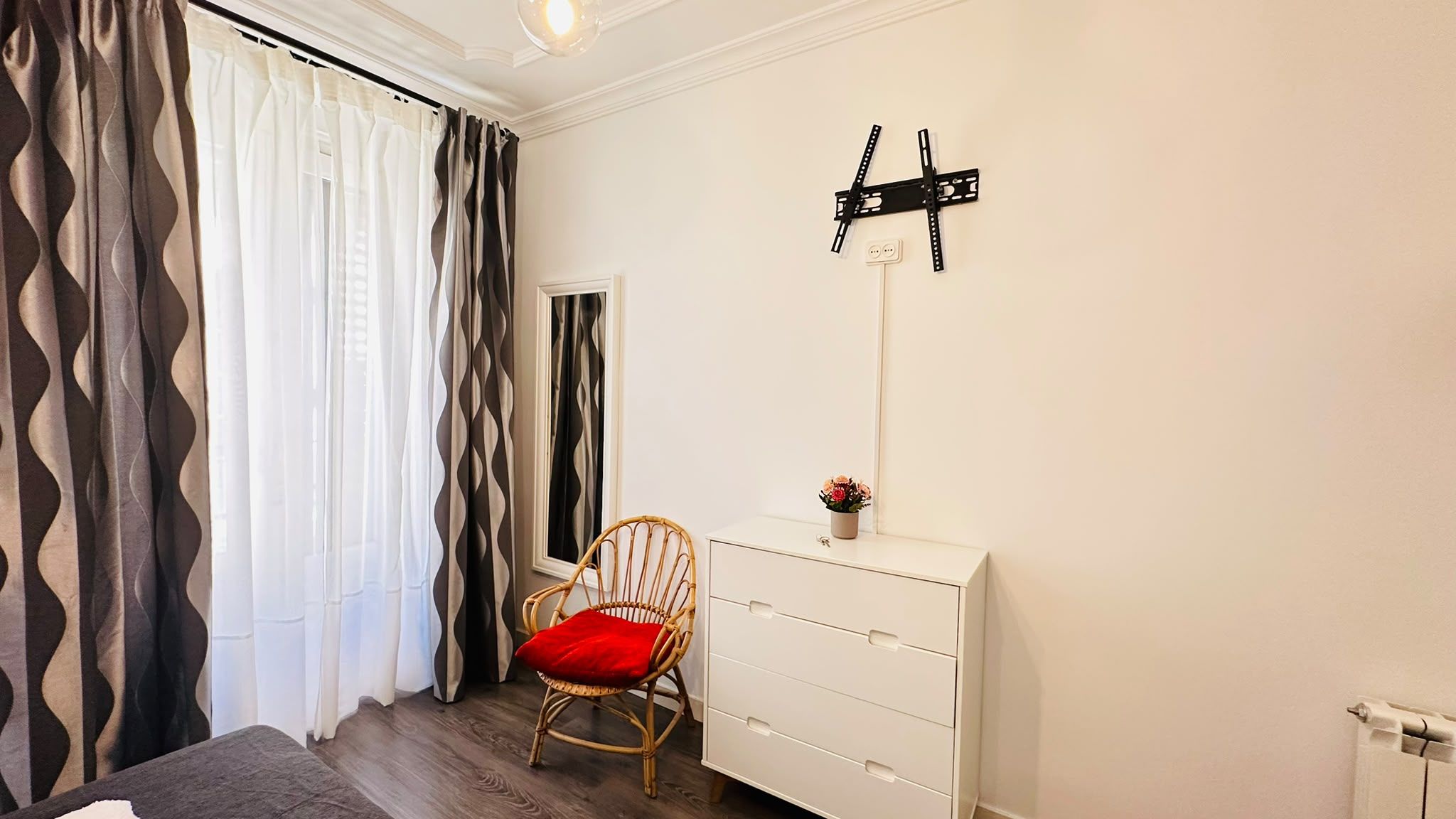 A bedroom with a bed, a chair, a dresser, a mirror, and a wall-mounted light fixture.