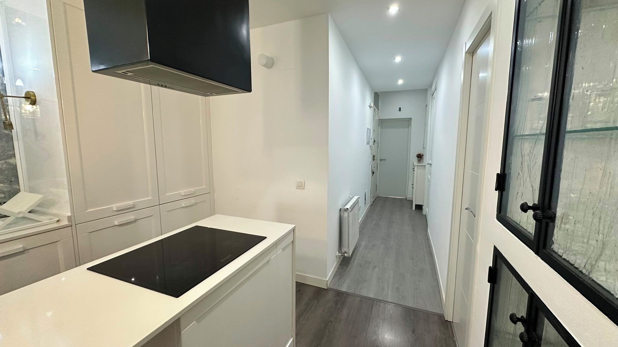 A narrow hallway with a kitchen area on one side, featuring white cabinetry and a black induction cooktop.