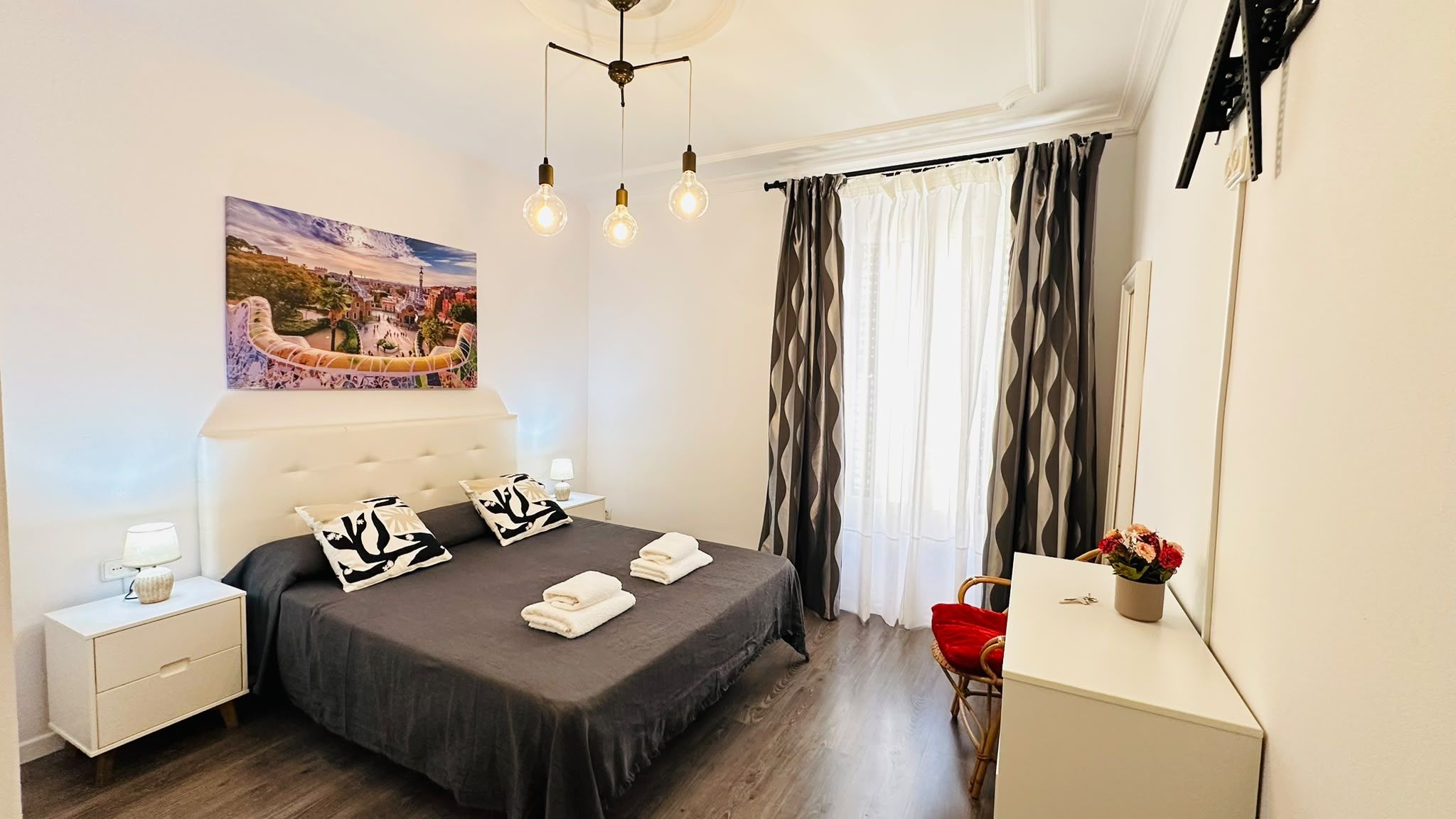 Un dormitorio con cama, mesitas de noche, escritorio, silla, un cuadro y una ventana con cortinas.