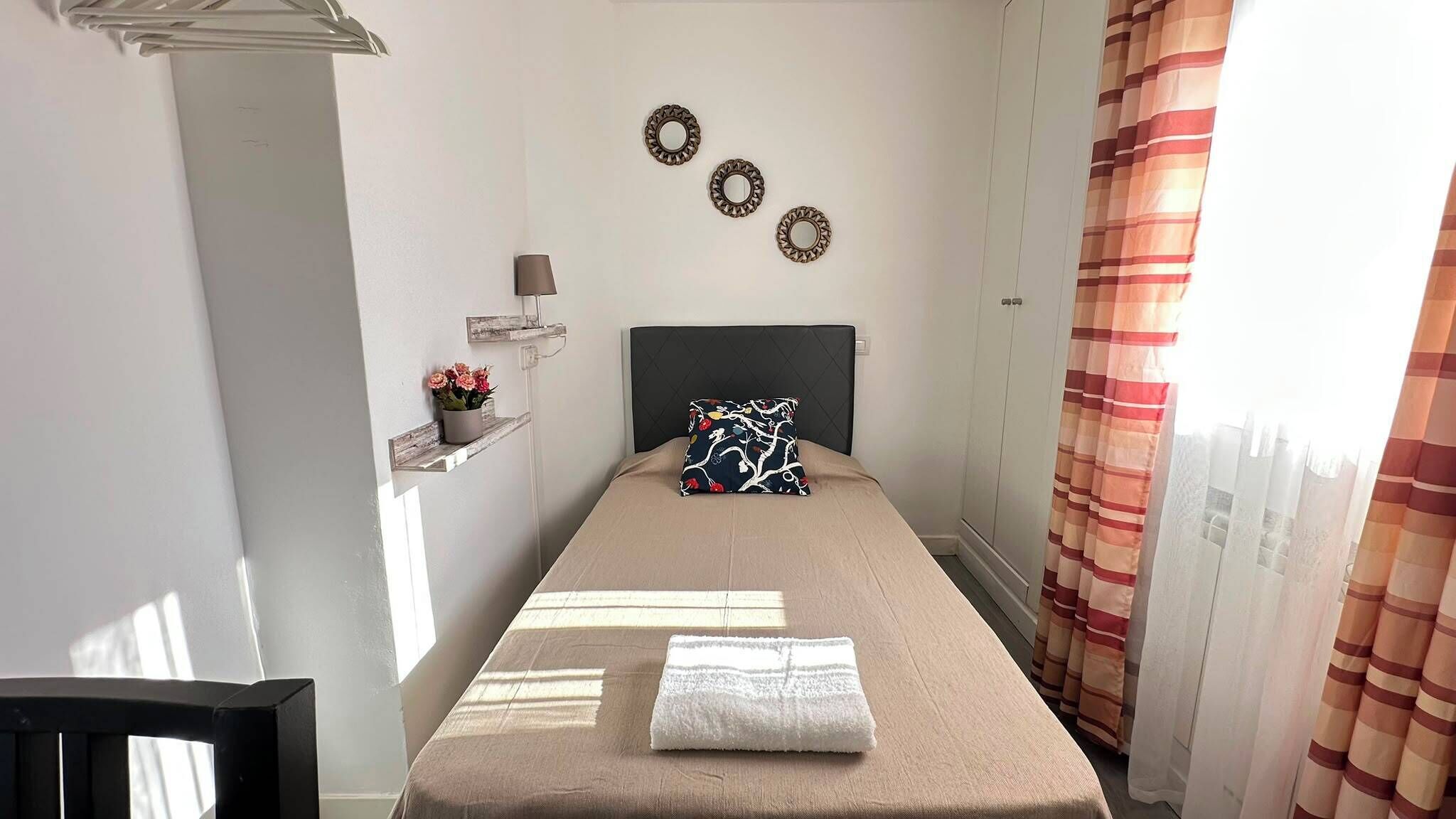 A narrow room with a bed, a small shelf with flowers, and a headboard with wall-mounted decorations.