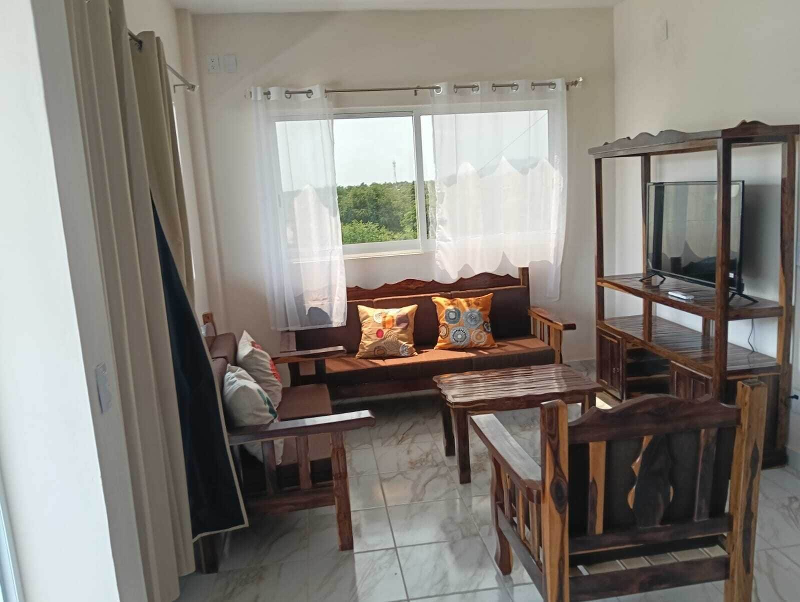 Een woonkamer met een bank, een televisie op een houten dressoir en een raam met dunne gordijnen.