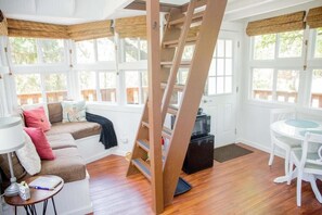 Una habitación acogedora con una escalera de madera, un sofá con cojines, una mesita con una lámpara y un comedor con sillas.