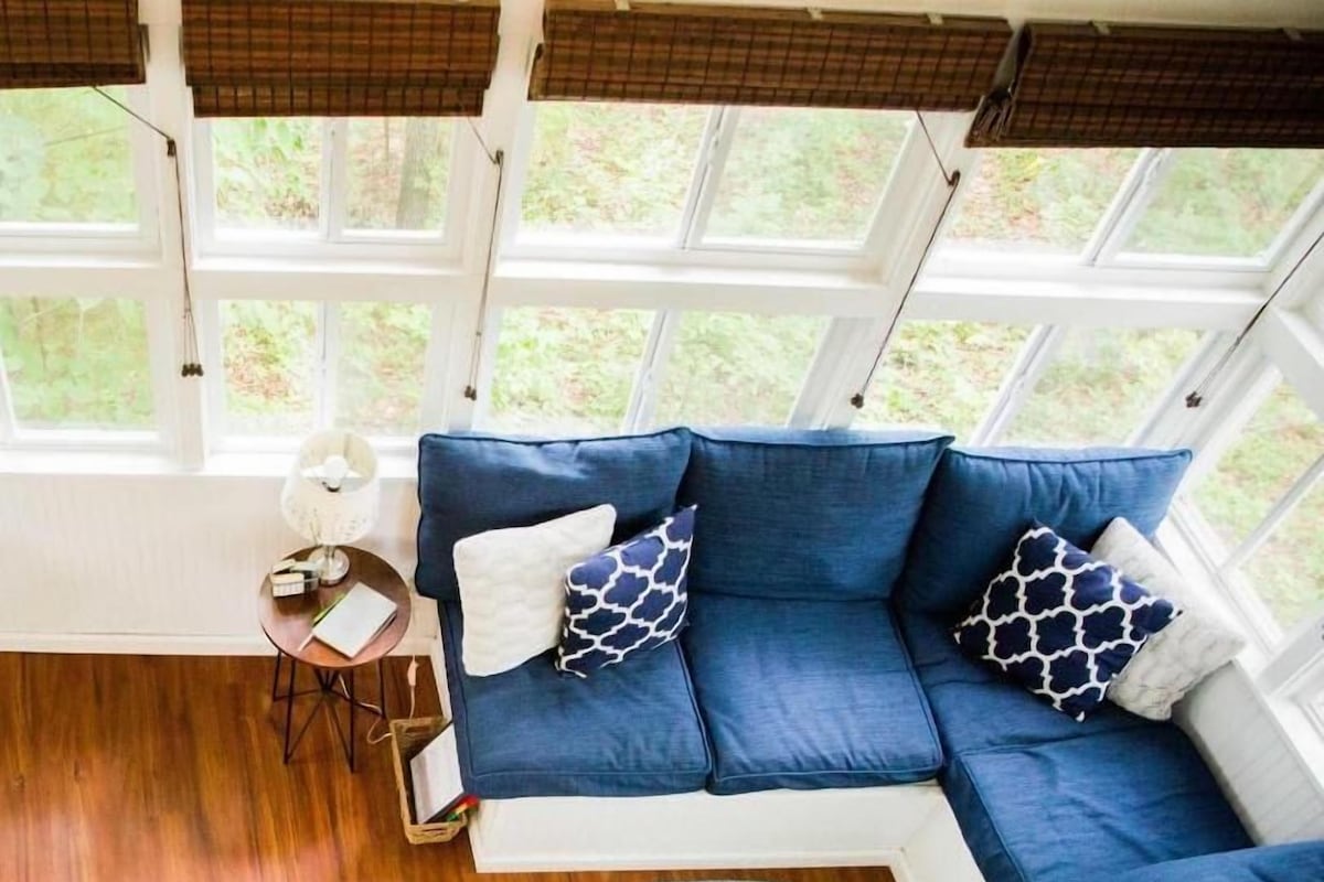 Una habitación con un sofá azul, un tapete redondo pequeño, una mesa con una lámpara y una ventana con vistas a zonas verdes.