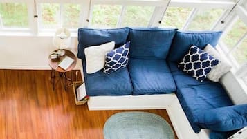 Una habitación con un sofá azul, un tapete redondo pequeño, una mesa con una lámpara y una ventana con vistas a zonas verdes.