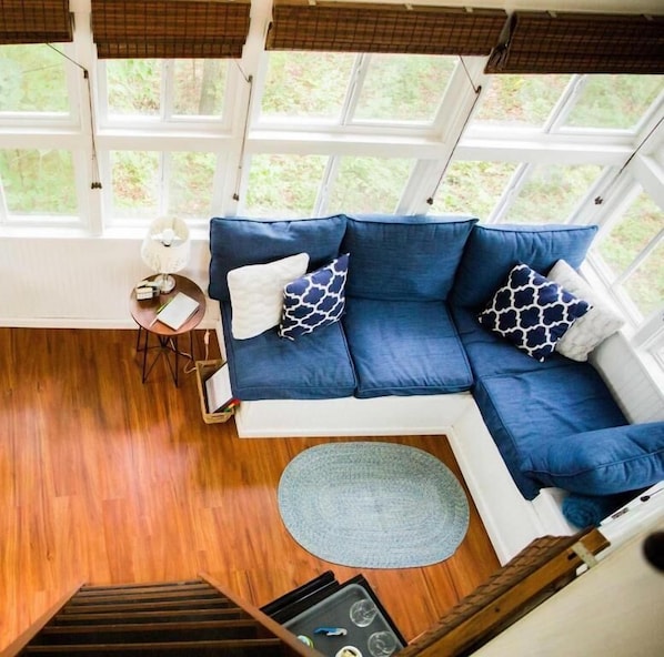 A room with a blue sofa, a small round rug, a table with a lamp, and a window with a view of greenery.