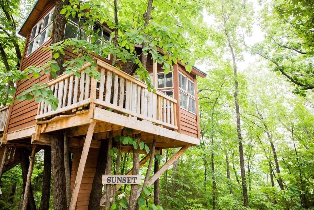 A treehouse with a wooden deck and a sign that reads 'SUNSET.'