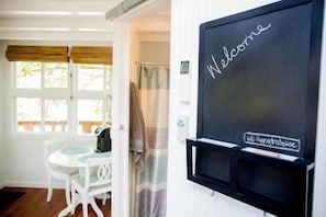 Una habitación con una puerta blanca, una ventana con persianas y una pizarra con la palabra 'Welcome' escrita, además de un letrero que indica 'Wifi: Sunrise Treehouse'.