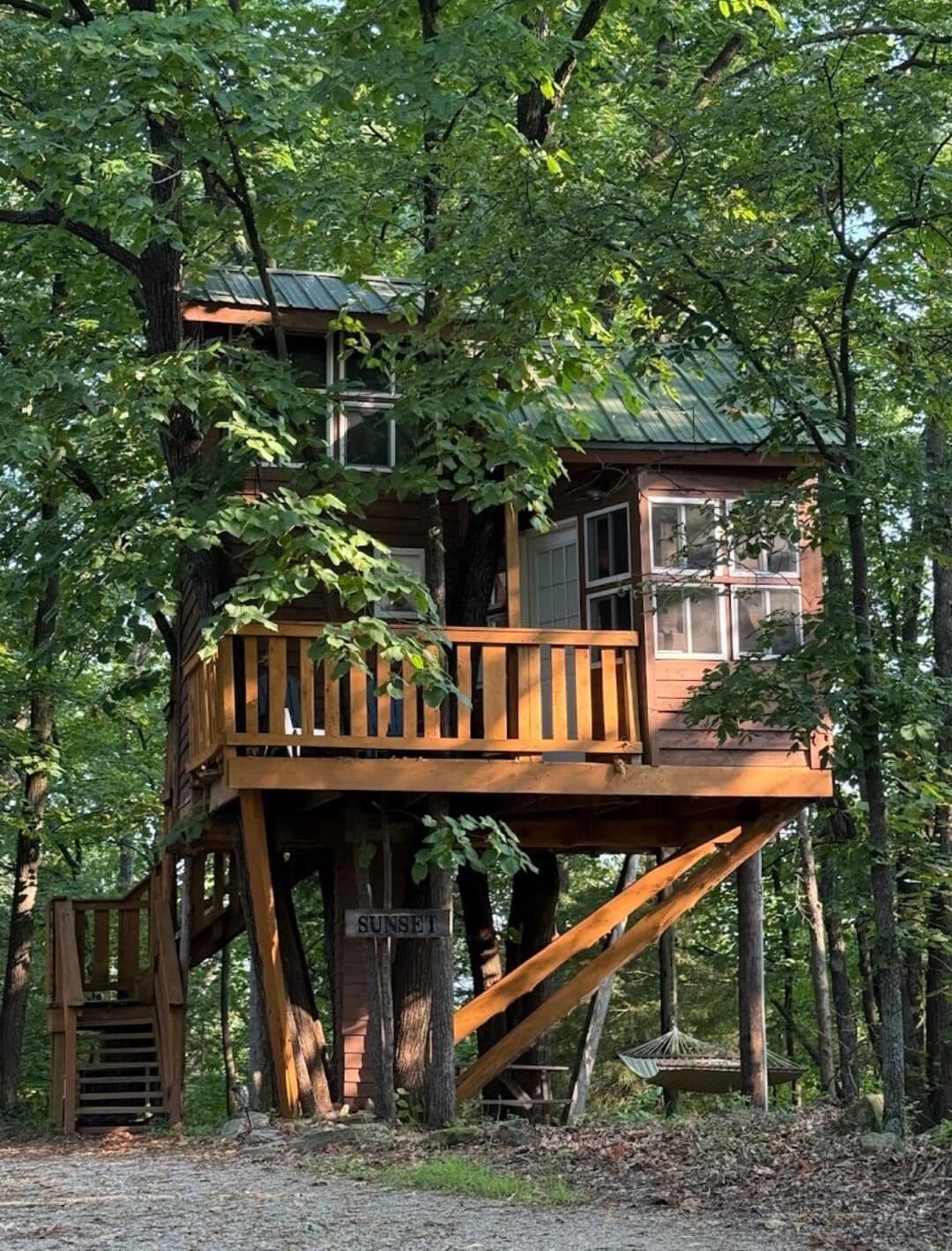 Ein Baumhaus mit einer Holzterrasse, einer Treppe und einem Schild mit der Aufschrift 'SUNSET'.
