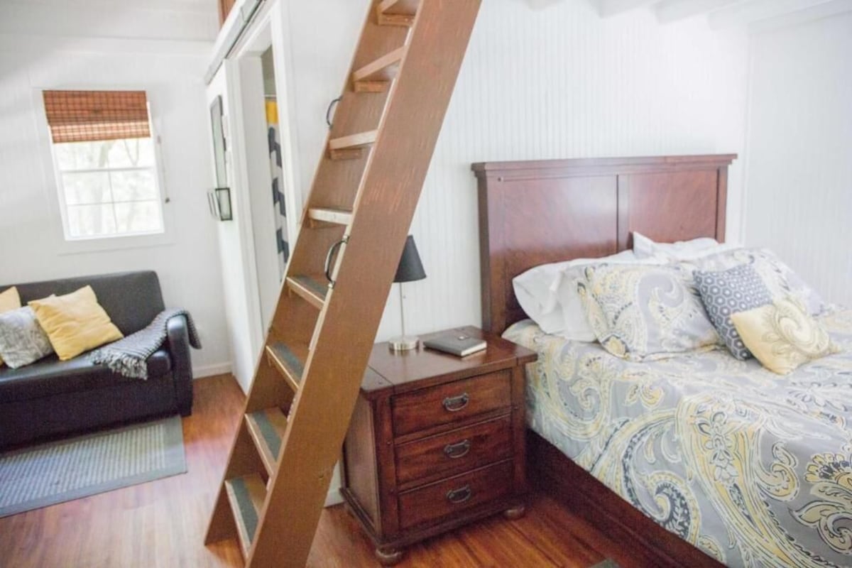 Un dormitorio con una escalera de madera, una cama con ropa de cama estampada, una mesita de noche, un aparador y un sofá de color oscuro.