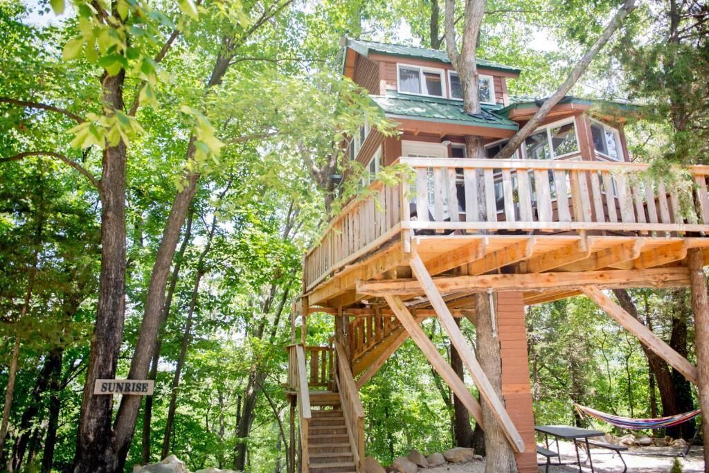 Uma casa na árvore com deck de madeira e escadas, rodeada por árvores.
