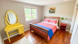 Un dormitorio con una cama de madera, un tocador amarillo con espejo, una ventana y una pequeña estantería roja.
