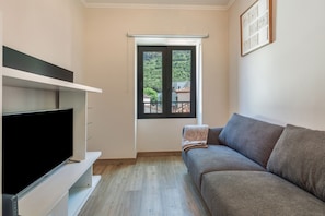 Ein modernes Wohnzimmer mit einem Flachbildfernseher, einem grauen Sofa und einem Fenster mit Blick auf Grünflächen.