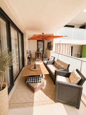 A balcony with a wooden table, wicker furniture, and a large orange umbrella. - New apartment, terrace & parking – Résidence Boréal, Montpellier (Monptellier)