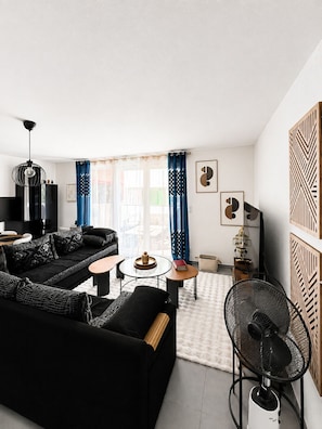 A modern living room with a black sofa, a round coffee table, and a black chair. There is a small round table with a book and a decorative item. The room has a white ceiling and walls, with a large window covered by blue curtains.