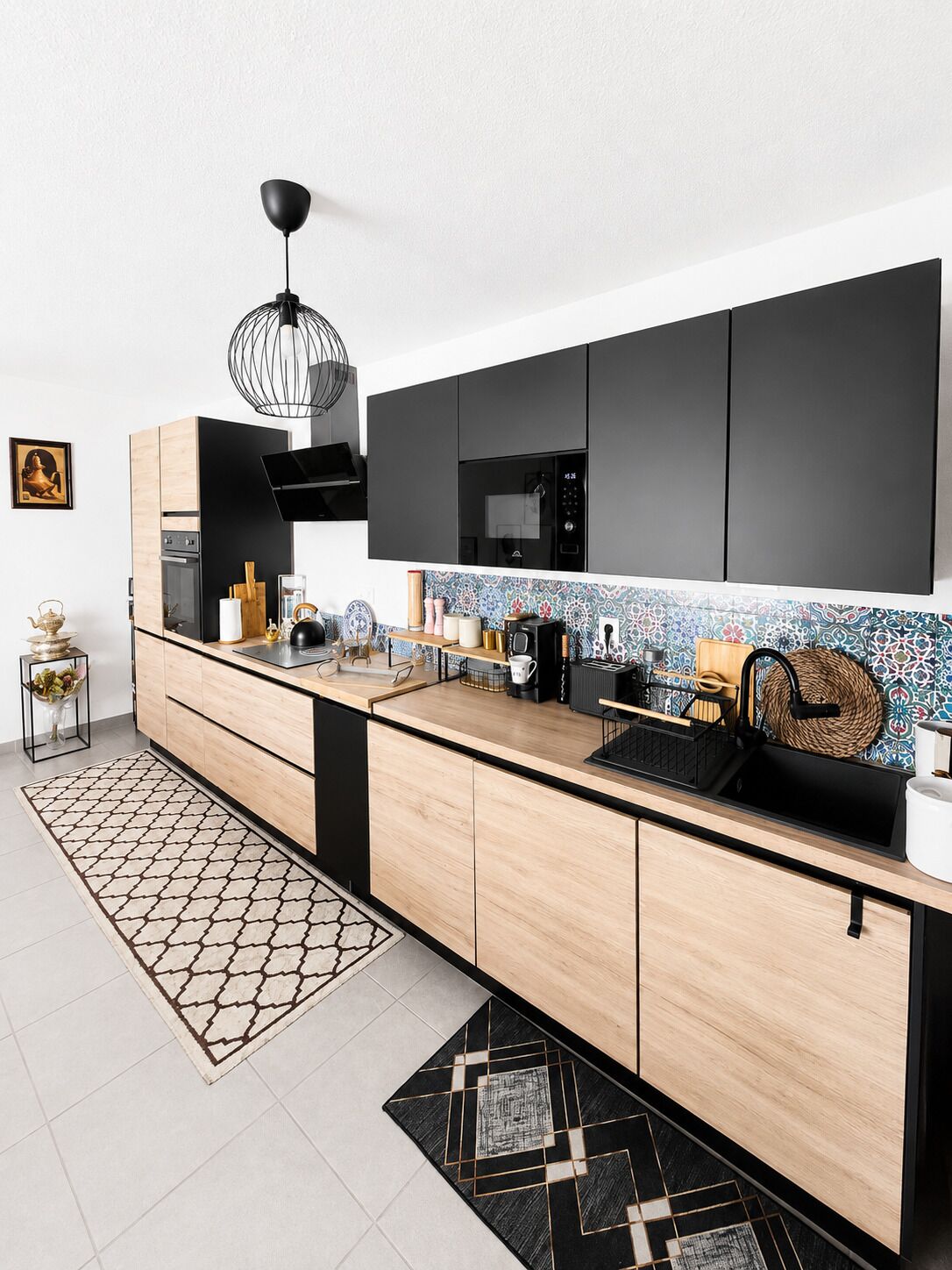 A modern kitchen with a black microwave, wooden cabinets, a patterned backsplash, and a hanging light fixture.