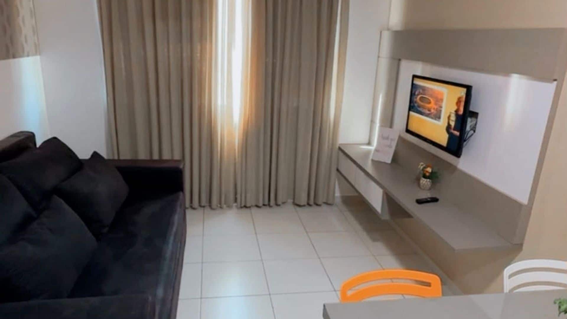 Olohuoneessa on tumma sohva, seinälle asennettu televisio, pieni pöytä, jossa on kaukosäädin, ja verhoilla peitetty ikkuna.