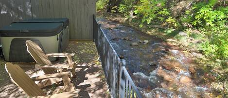 A wooden deck with a metal railing overlooking a stream, a wooden chair, and a covered seating area.