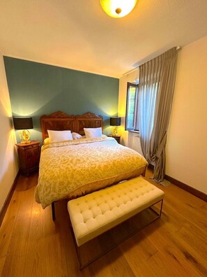 A bedroom with a bed, bedside lamps, a bench, and a window with curtains. - The Old Village (Vignola)