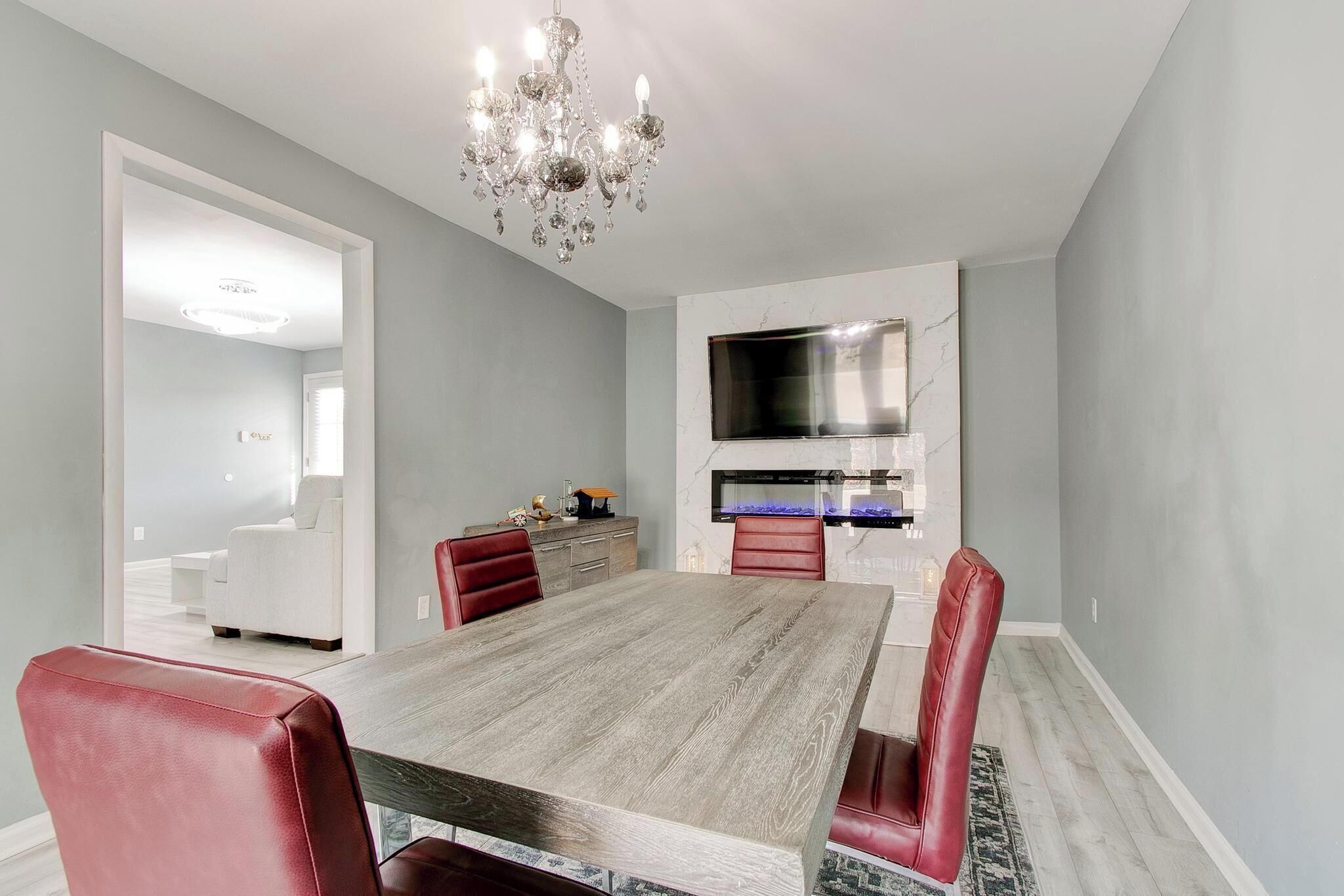 A dining room with a long table, red chairs, a chandelier, a kitchen area with a TV and stove, and a white sofa in the adjacent room.