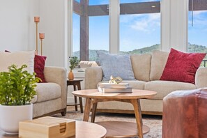 A modern living room with a sofa, a coffee table, and a view of a green landscape through large windows.