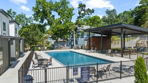A swimming pool with lounge chairs and a covered seating area, surrounded by a fence and trees.