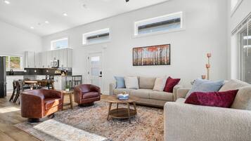 A spacious living area with a sofa, armchair, and coffee table, a kitchen in the background, and a painting on the wall.