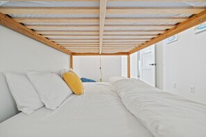 A single bed with white bedding and a yellow pillow in a room with white walls and a wooden ceiling structure.