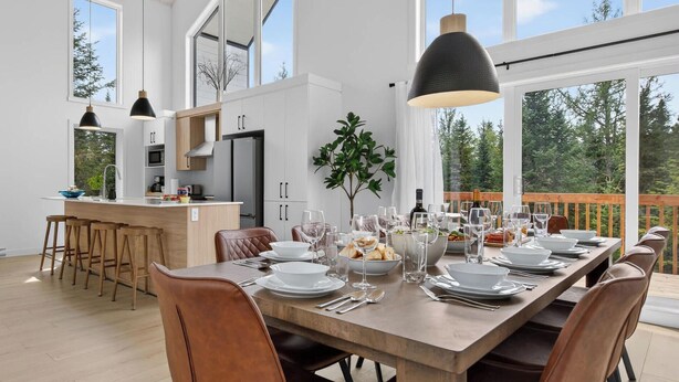 A modern dining area with a wooden table set for a meal, large windows, and a view of the outdoors.