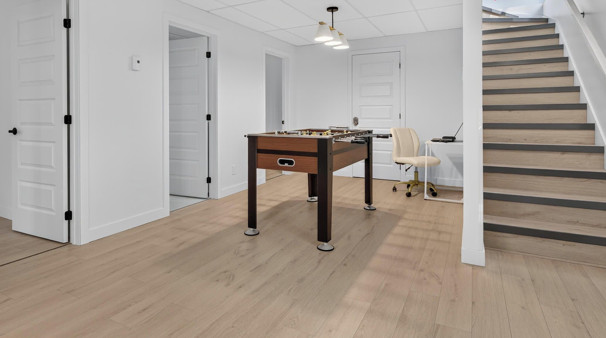 Une pièce avec un baby-foot, un bureau avec un ordinateur et une chaise. Un escalier mène à un autre niveau.