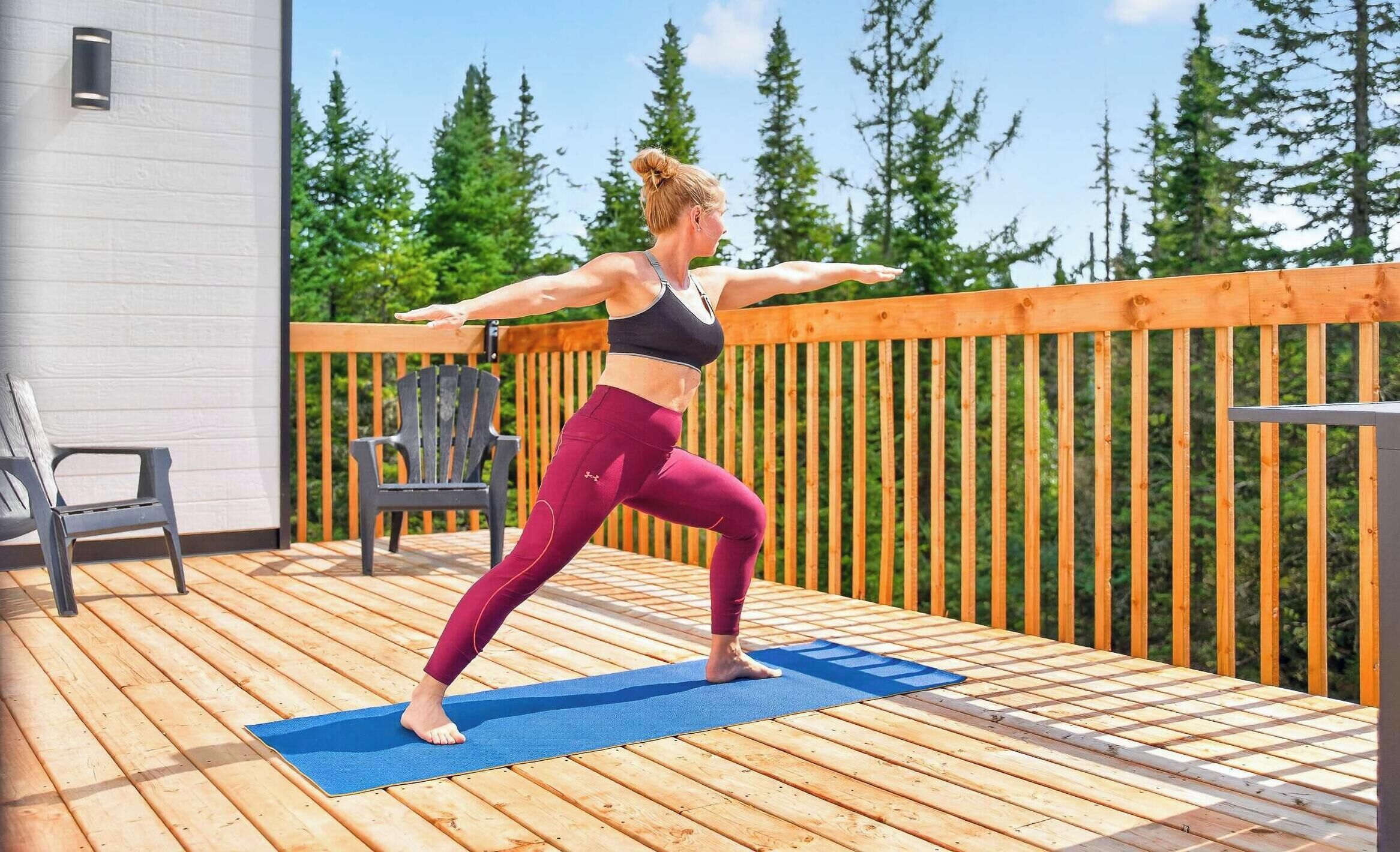 A person performing a yoga pose on a wooden deck with a blue mat, surrounded by a wooden fence and chairs.