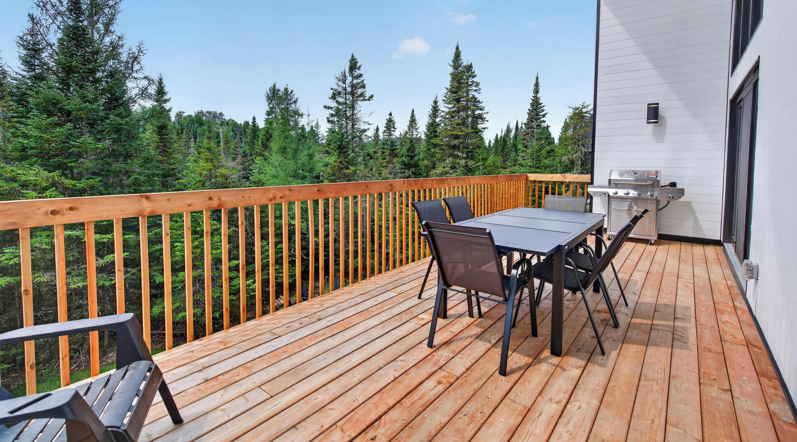 Una terraza de madera con mesa y sillas, una parrilla y vistas a un bosque.