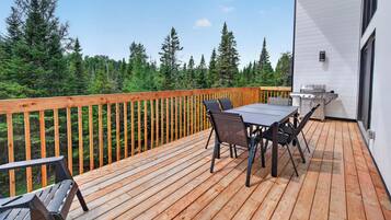 Una terraza de madera con mesa y sillas, una parrilla y vistas a un bosque.