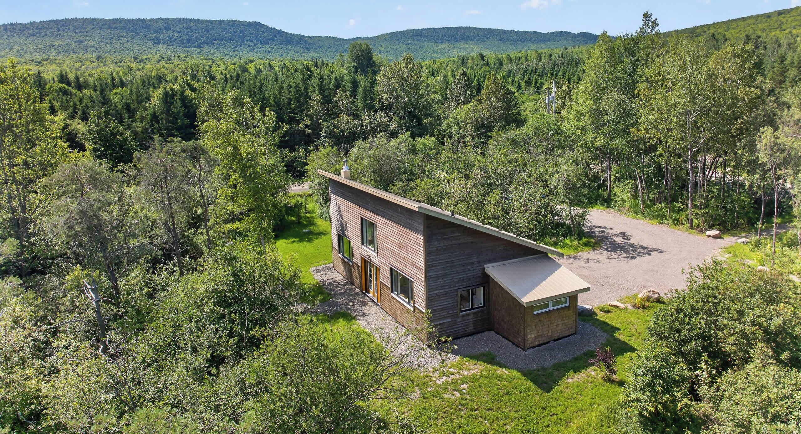 A modern two-story house with a brick exterior, surrounded by dense forest and hills.