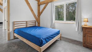 A bedroom with a wooden bed frame, a single bed with a blue mattress, a window with a view of trees, and a wooden nightstand with a lamp. - The Enlightened - Retreat Under the Megantic Stars (Notre-Dame-des-Bois)