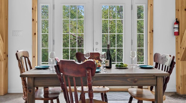 A dining area with a wooden table and chairs, a fire extinguisher on the wall, and a view of trees through large windows.