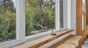 A room with a large window overlooking a garden, a wooden bench, and a small table with a bottle.