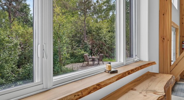 A room with a large window overlooking a garden, a wooden bench, and a small table with a bottle.