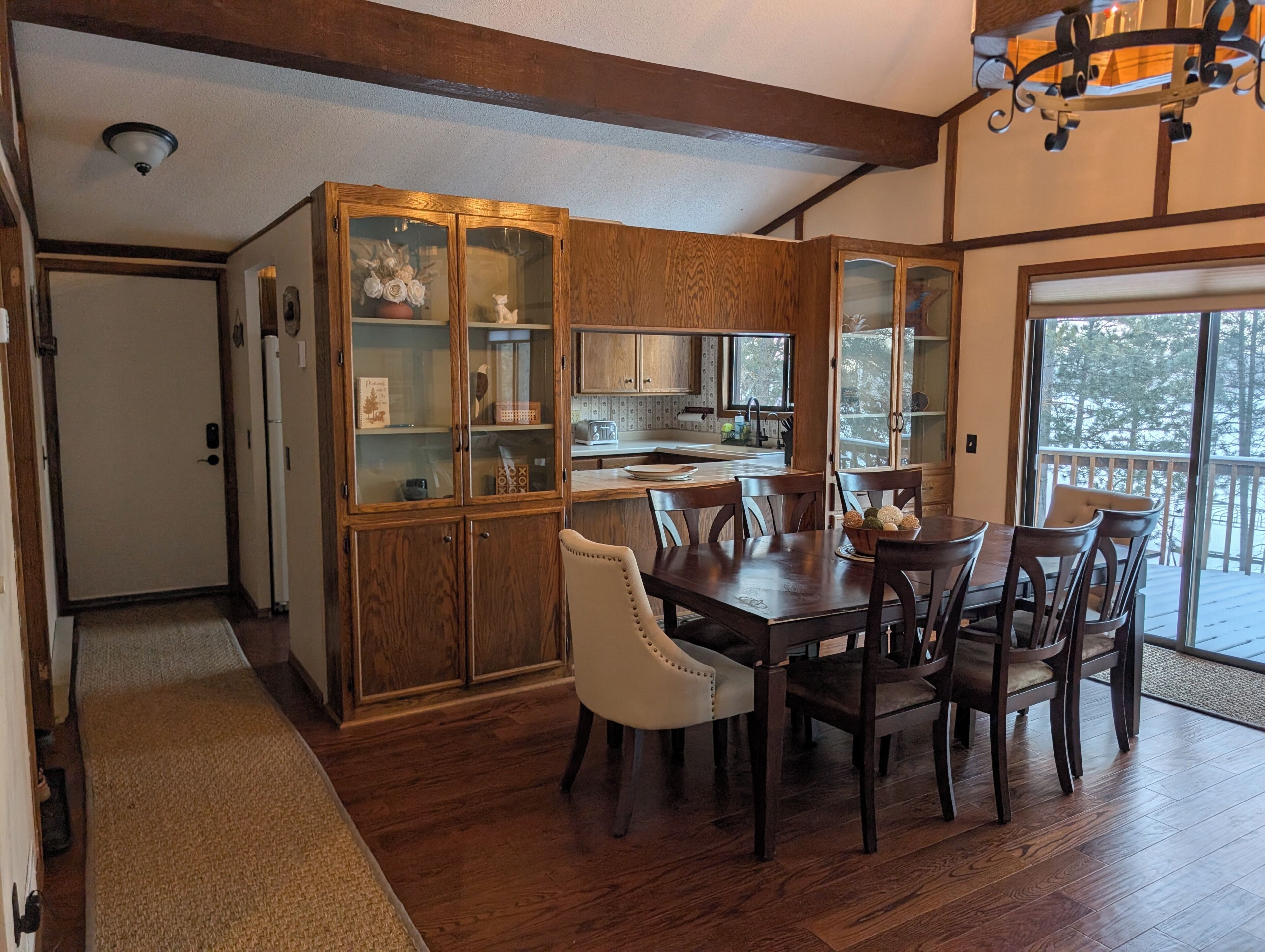 A dining area with a wooden table and chairs, a kitchen with wooden cabinets, and a view of the outdoors through a glass door.