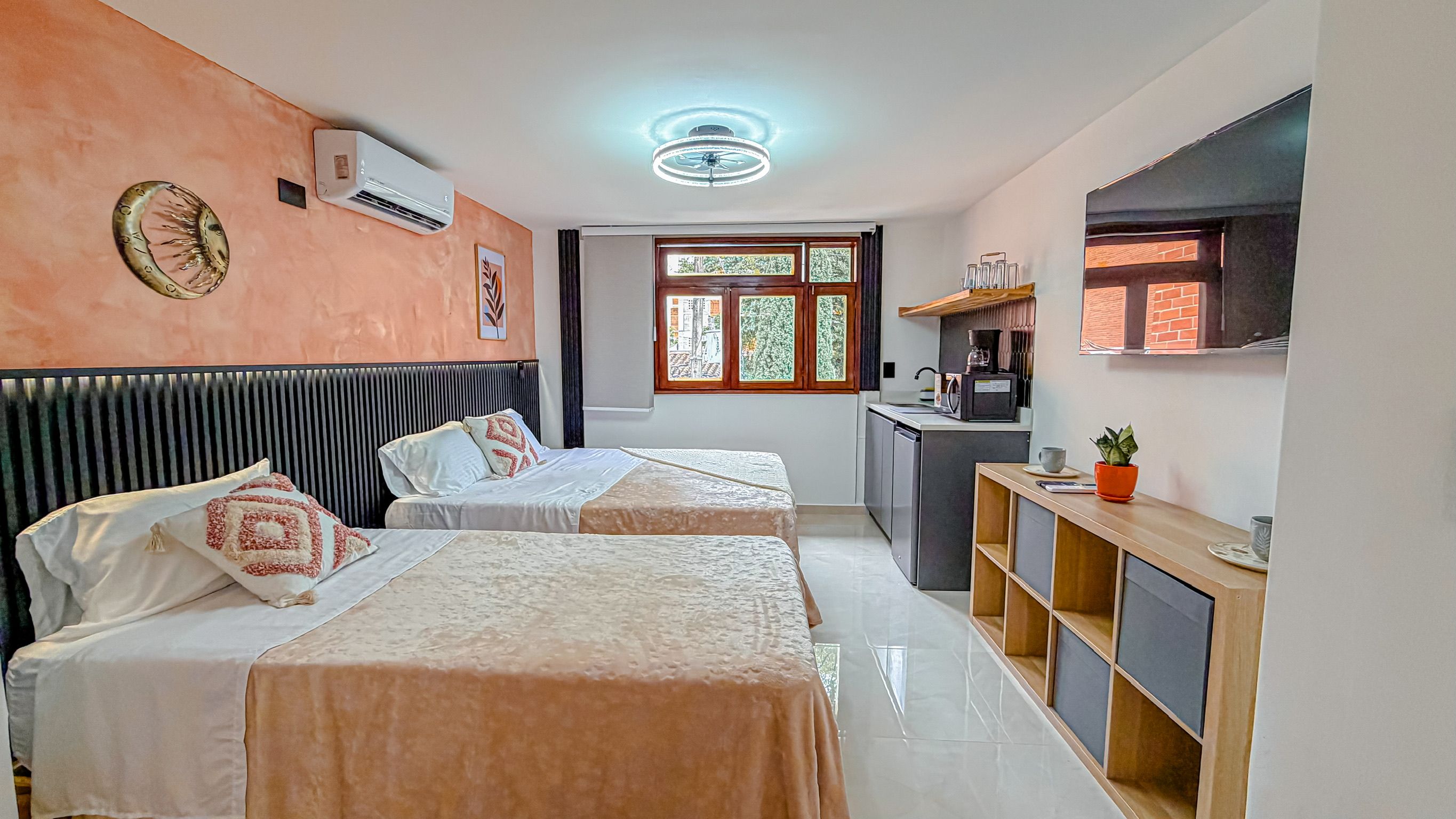 A hotel room with two beds, a kitchenette, a window with a view, and a ceiling fan.