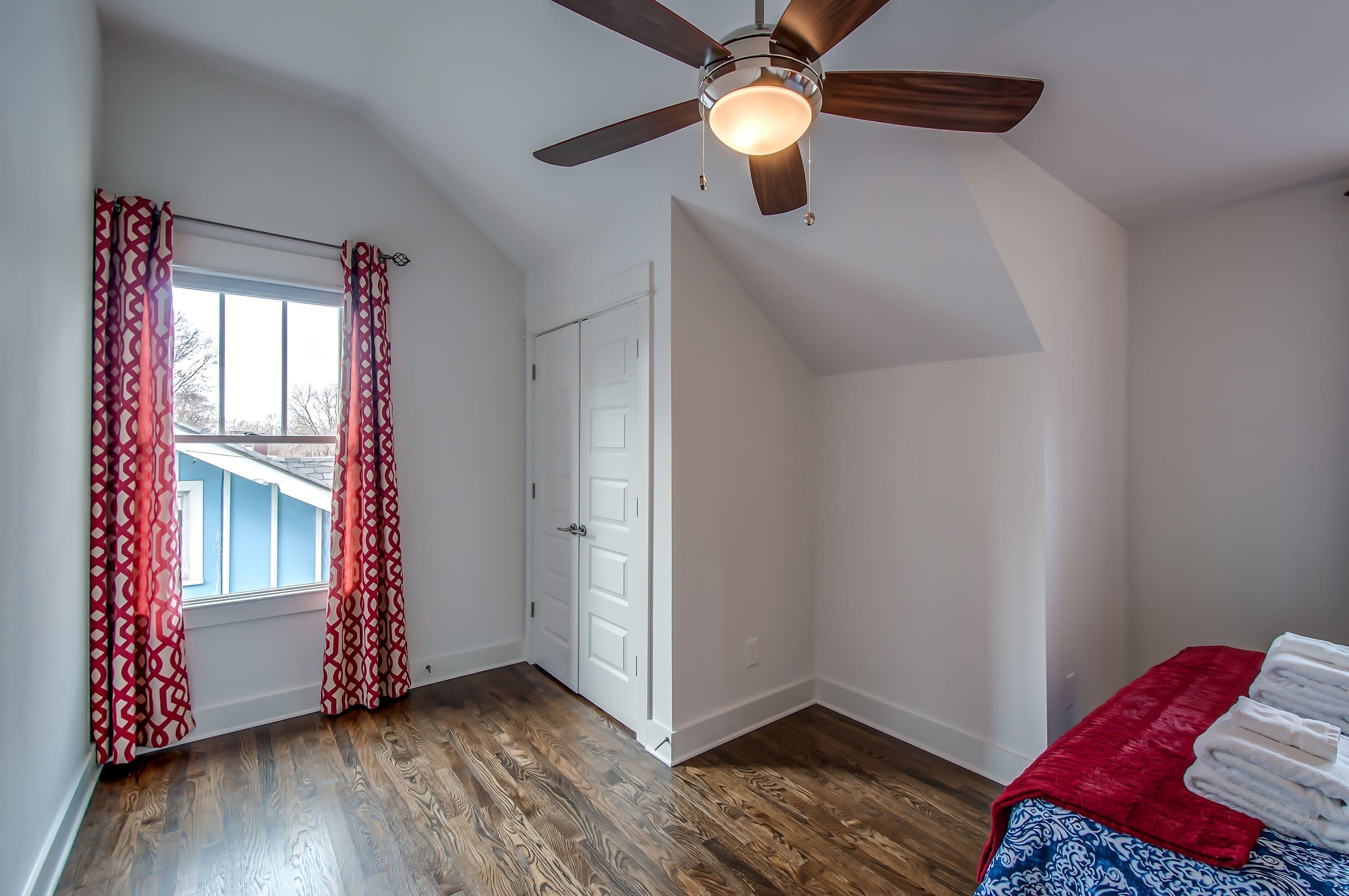 A room with a sloped ceiling, a ceiling fan, a window with patterned curtains, a white door, and a bed with a red blanket.