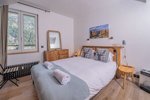 A bedroom with a bed, a dresser, a radiator, a window with a view, a painting, a lamp, and a table. - Majestic Panorama - Stunning Mont Blanc Views|Fireplace|Central Chamonix (Chamonix-Mont-Blanc)
