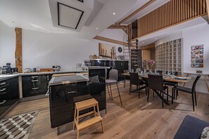 A modern kitchen with a central island, a dining area with a wooden table, and a loft space with a wooden staircase. - Majestic Panorama - Stunning Mont Blanc Views|Fireplace|Central Chamonix (Chamonix-Mont-Blanc)