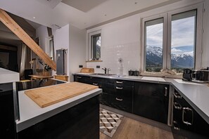 A modern kitchen with a wooden cutting board, black cabinets, and a view of snow-capped mountains through large windows. - Majestic Panorama - Stunning Mont Blanc Views|Fireplace|Central Chamonix (Chamonix-Mont-Blanc)
