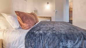 Quarto com cama, travesseiros, luminária na parede e porta para outro cômodo.