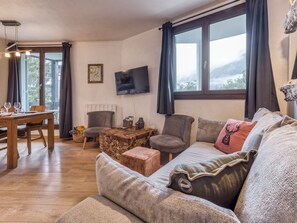 Una sala de estar acogedora con una mesa de comedor de madera, asientos cómodos, un televisor de pantalla plana y ventanas grandes con cortinas.